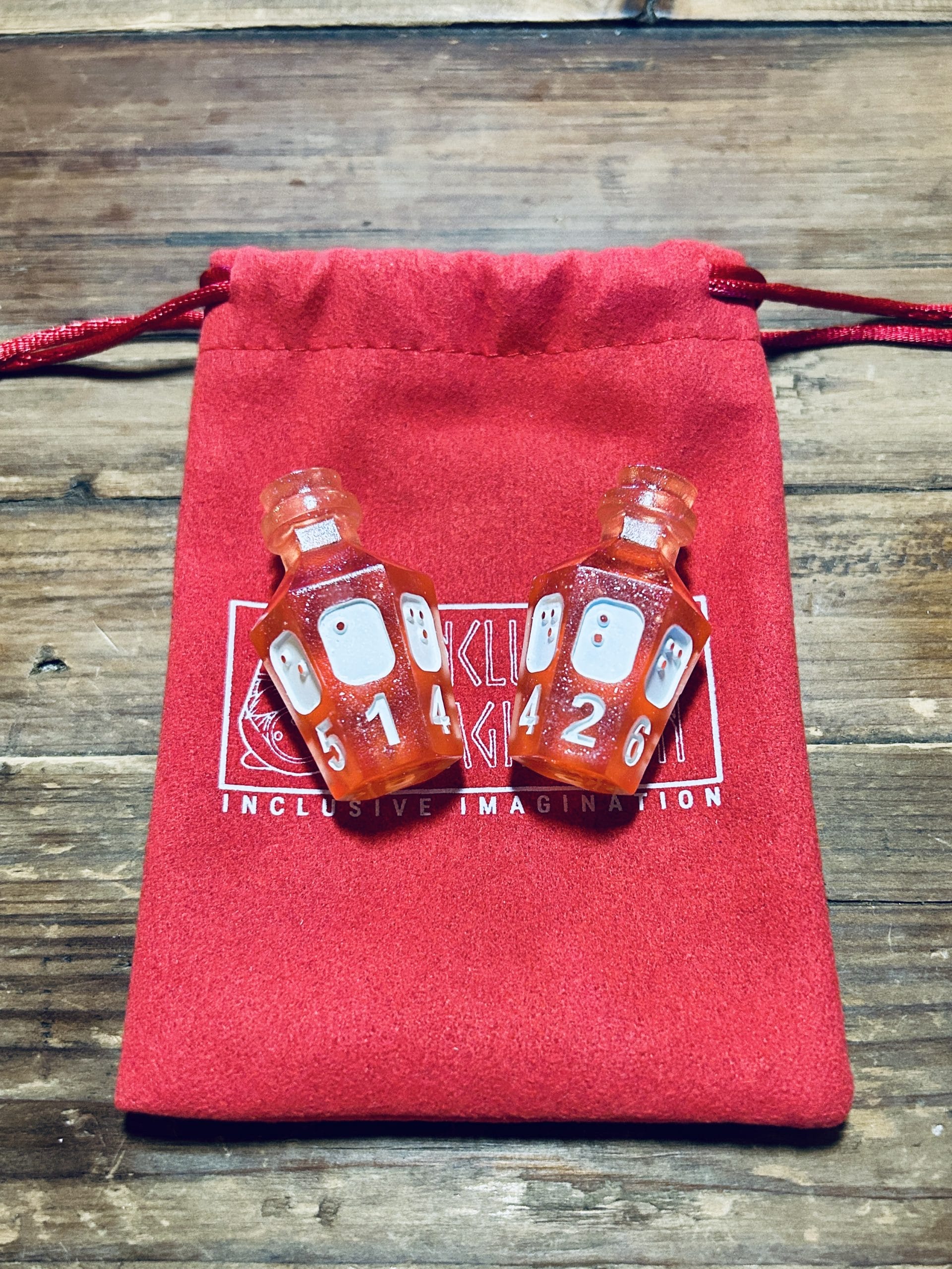 2 Rose Glitter potion D6s lay on a red microfiber pouch with inclusive imagination logo. The d6s show numbers 1 and 2 in both print and braille.