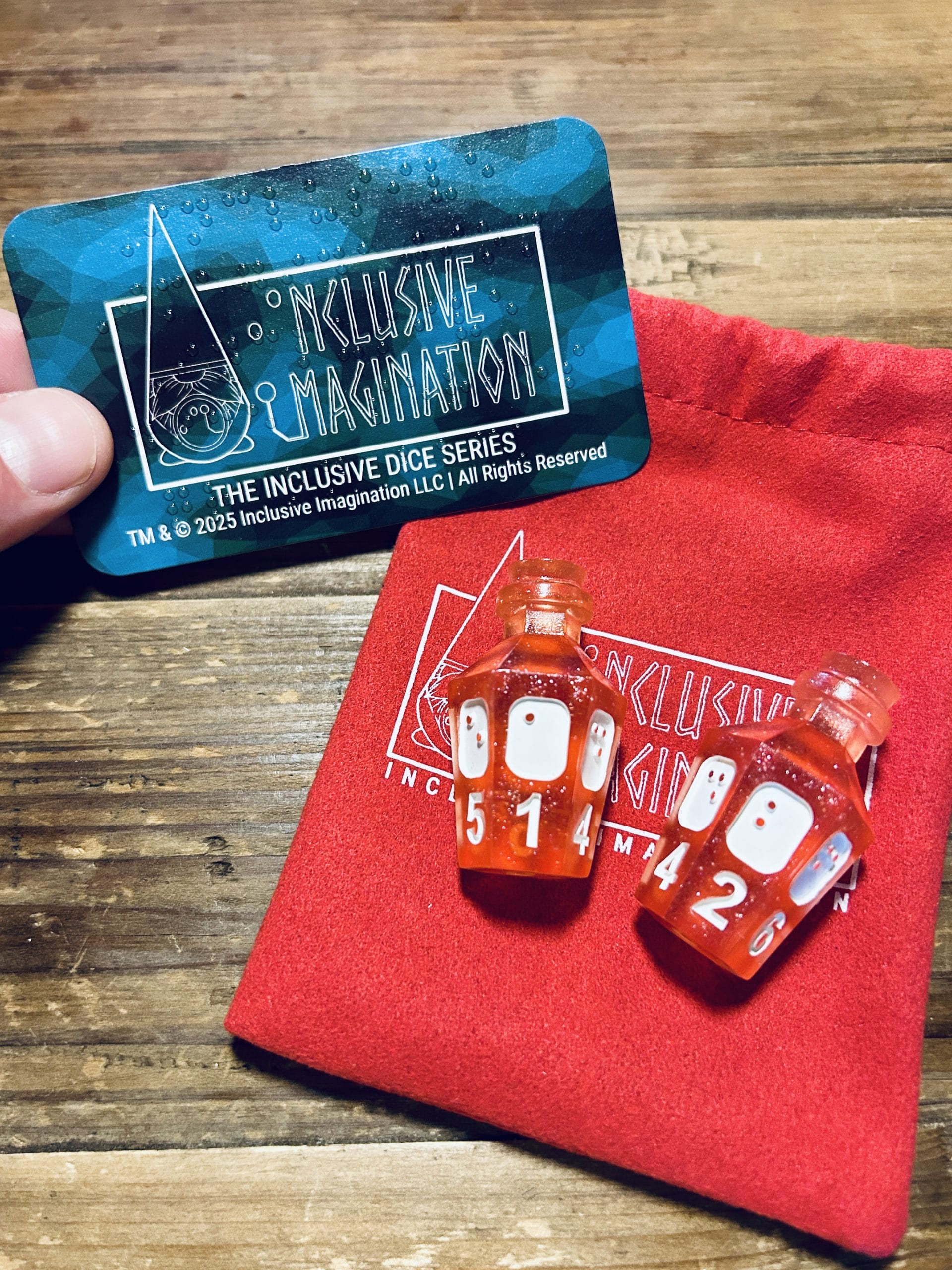 2 Rose Glitter potion D6s lay on a red microfiber pouch with inclusive imagination logo. An accessible card insert is shown above with print and braille.
