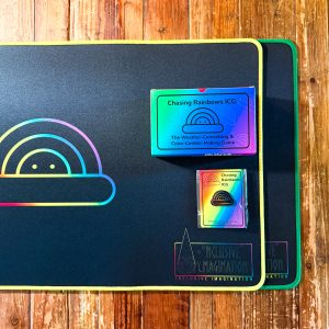 1 Chasing Rainbows Game, 1 Rainbow Logo Pin, 1 Yellow Playmat, and 1 Green Playmat on a Table