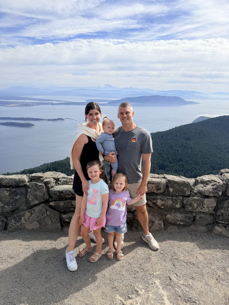 A photo of Garrett, his wife, Lexi, and 3 daughters (ages 5, 3, and 1) on top of Mt. Constitution in Orcas Island overlooking the San Juan Islands in the distant background. 